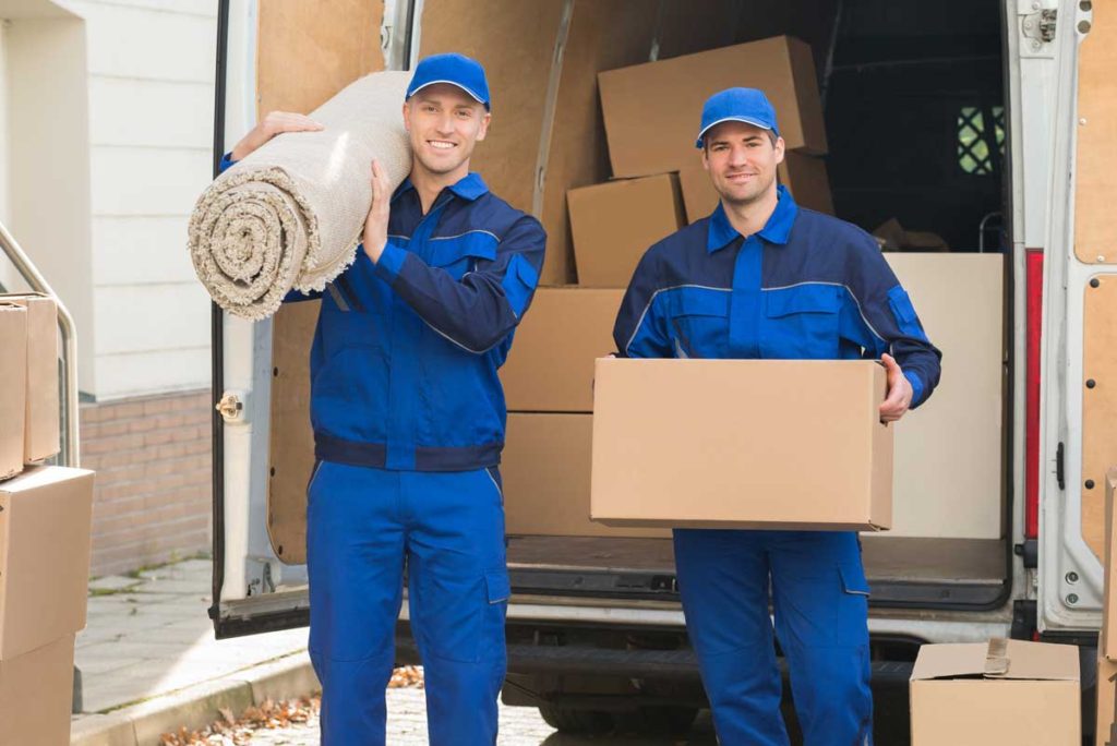 two men in blue uniforms holding a box and a carpet in front of open truck